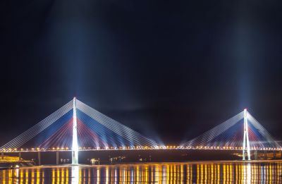 мост на остров Русский
В январе 2013 года завершился грандиозный проект художественного освещения моста на остров Русский во Владивостоке, который был построен в рамках подготовки города к саммиту АТЭС-2012. Самый высокий и самый длинный в мире вантовый мост соединил круглогодичной переправой материковую и островную части Владивостока, став ключевым объектом транспортной инфраструктуры Дальневосточного региона. 

Подсветка мостового перехода на остров Русский выполнена по уникальному проекту специалистов компании "МТ Электро". Основная идея концепции архитектурно-художественного освещения – создание иллюзии парящего в воздухе моста при активной подсветке опор и множества вант, окрашенных в цвета российского флага. 

Безусловно, проектирование декоративной подсветки мостов – особое направление светового дизайна, требующее повышенного внимания к характерным функциональным и архитектурным особенностям данных сооружений. Подсветка не должна влиять на ночное освещение проезжей части, выделяться на конструкциях моста в дневное время, световой поток не должен слепить водителей наземного транспорта и судоводителей. Прежде всего, это влияет на выбор оборудования, определение тех или иных приемов подсветки, выбор цвета и интенсивности каждого источника света. Сооружение такого колоссального масштаба, как мост на остров Русский, потребовало применения инновационных инженерных решений. Ведь помимо создания максимально комфортной и безопасной световой среды, необходимо было учитывать сложные климатические условия, такие как ветровые нагрузки до 51 м/cек на высоте 70 м, сейсмическая активность до 8,1 баллов по шкале MSK-64, перепады температуры от −31°С до +37°С. Оптимальным вариантом было признано светодиодное освещение: для линейной подсветки основного пролетного строения были выбраны светодиодные светильники специализированной серии IntiLINE marine производства "IntiLED". Усиленное климатическое исполнение топового изделия модельного ряда "IntiLED" отличается высокой степенью защиты от внешних возде
Keywords: Светодиодное освещение, светодиодная подсветка, архитектурное освещение, архитектурная подсветка, светодиодные светильинки, светодиодное оборудование 